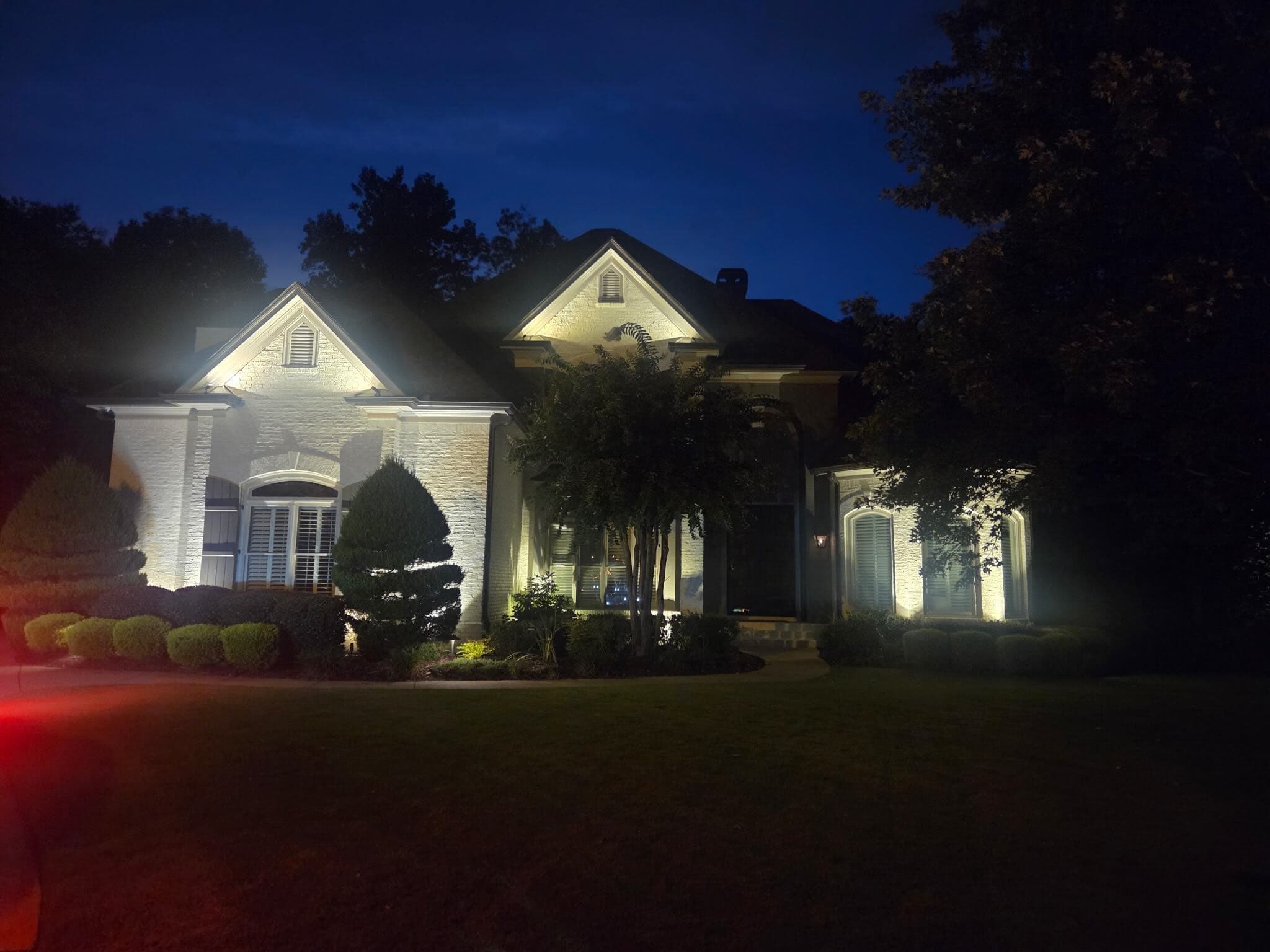 White-brick home in Dacula, GA 30019 lit at night with warm gable and facade uplighting and a mature crape myrtle glowing at the center of the front yard