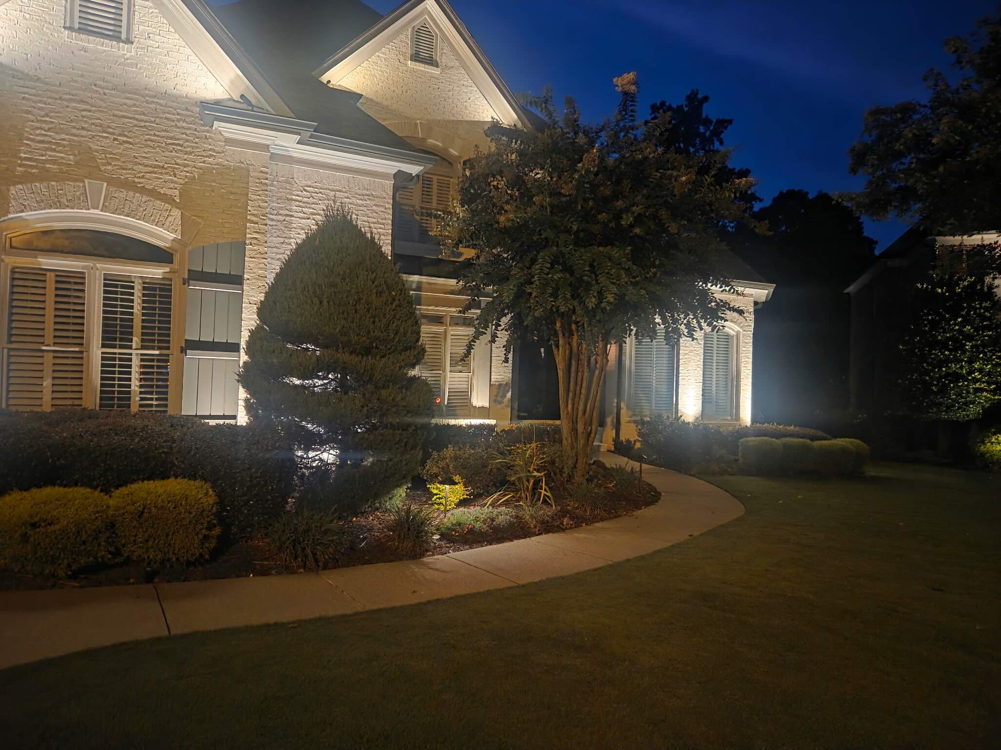 Angled view of a white-brick home in Dacula, GA 30019 with a curved walkway lit by path lights, a spiral topiary, and warm uplighting on the facade and windows