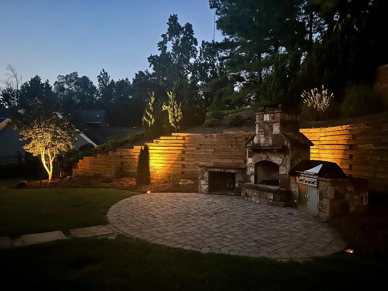 Outdoor fireplace, grill, and paver patio lit with layered warm lighting at night